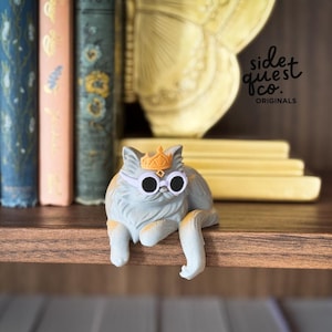 May include: A light gray cat figurine wearing white-rimmed sunglasses and a gold crown. The cat is lying down with its front paws hanging over the edge of a wooden shelf. The text "side quest co. ORIGINALS" is visible.