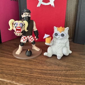 May include: A figurine of a dark-haired character wearing a black mask, brown vest, and white shorts with red hearts, holding a blonde-haired head, standing next to a white cat figurine wearing a gold crown and white sunglasses holding a drink.