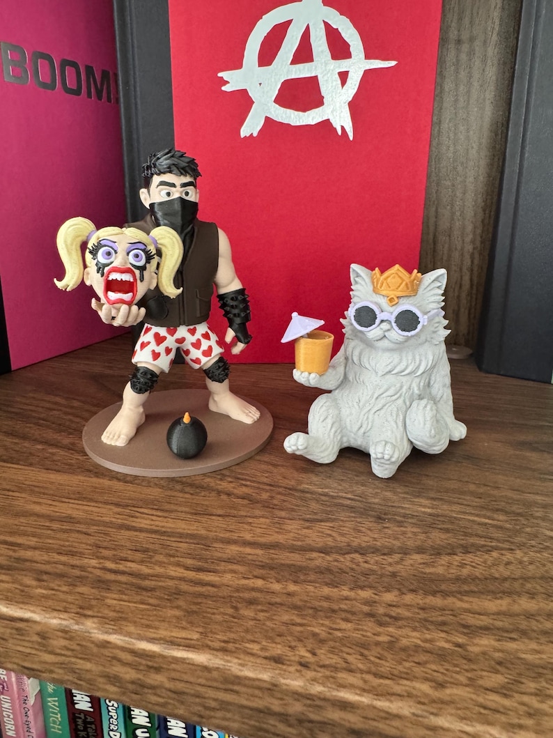 May include: A figurine of a dark-haired character wearing a black mask, brown vest, and white shorts with red hearts, holding a blonde-haired head, standing next to a white cat figurine wearing a gold crown and white sunglasses holding a drink.