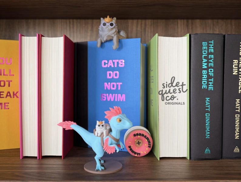 May include: A bookshelf displays several books with colorful spines. A blue book with the text "CATS DO NOT SWIM" has a cat figurine on top. A blue dinosaur figurine with a cat on its back is in front of the books. A pink donut-shaped object is also in front of the books.