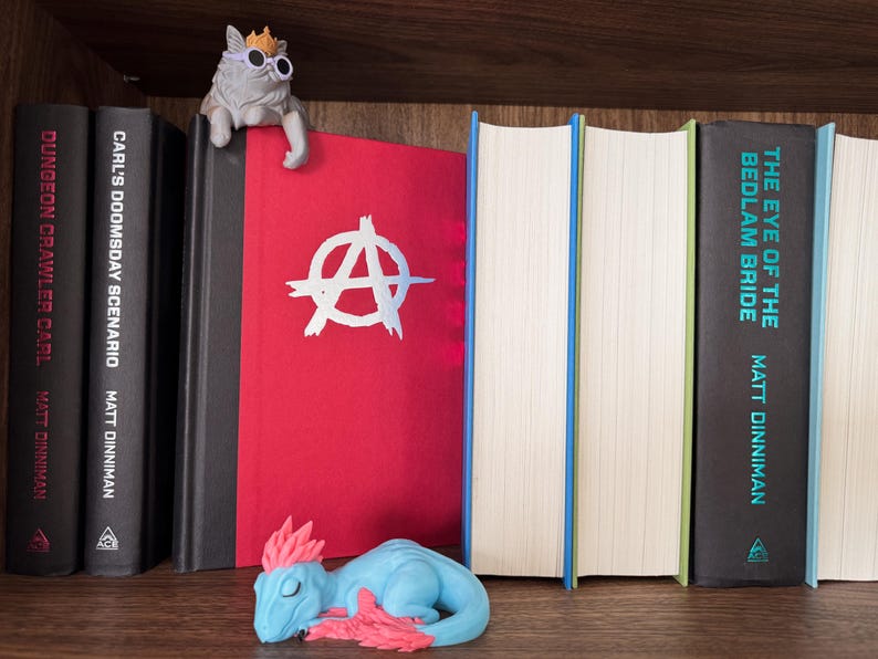 May include: A bookshelf containing books titled Dungeon Crawler Carl, Carl's Doomsday Scenario, and The Eye of the Bedlam Bride by Matt Dinniman. A red book with a white anarchy symbol sits between them, topped by a cat figurine wearing sunglasses.