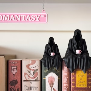 Może przedstawiać: R&oacute;żowy znak z napisem "ROMANTASY RD" wisi nad p&oacute;łką z książkami. Dwie czarne postacie w szatach siedzą na książkach, każda trzymając r&oacute;żowy kubek. Książki mają kolorowe okładki z tytułami.