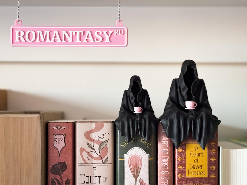 Może przedstawiać: R&oacute;żowy znak z napisem "ROMANTASY RD" wisi nad p&oacute;łką z książkami. Dwie czarne postacie w szatach siedzą na książkach, każda trzymając r&oacute;żowy kubek. Książki mają kolorowe okładki z tytułami.