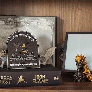 Könnte beinhalten: Ein schwarzes und goldenes Lesezeichen mit dem Text "Ich hatte die Zeit meines Lebens, Drachen mit dir zu bekämpfen". Das Lesezeichen ist in Form eines geöffneten Buches mit einem Drachen und Sternen auf dem Einband gestaltet. Das Lesezeichen liegt auf einem Buch mit dem Titel "Iron Flame" von Rebecca Yarros.