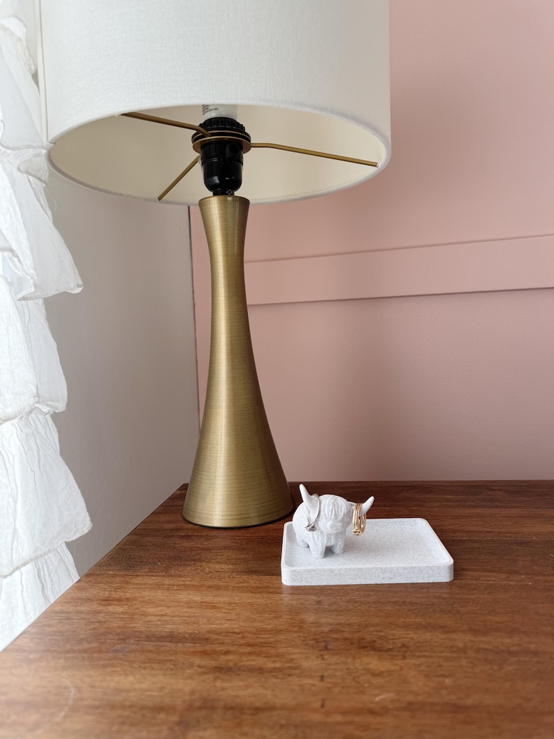 Op de afbeelding: Een witte keramische ringhouder in de vorm van een Schotse hooglander met gouden ringen aan zijn hoorns. De houder staat op een grijs, getextureerd vierkant dienblad. Het dienblad staat op een houten oppervlak naast een gouden tafellamp met een witte lampenkap.