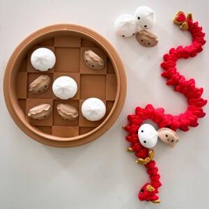 May include: A red dragon toy with a heart on its head, made of a soft, flexible material. The dragon is curled up around three white and brown steamed bun toys. The buns are also in a brown bamboo steamer.