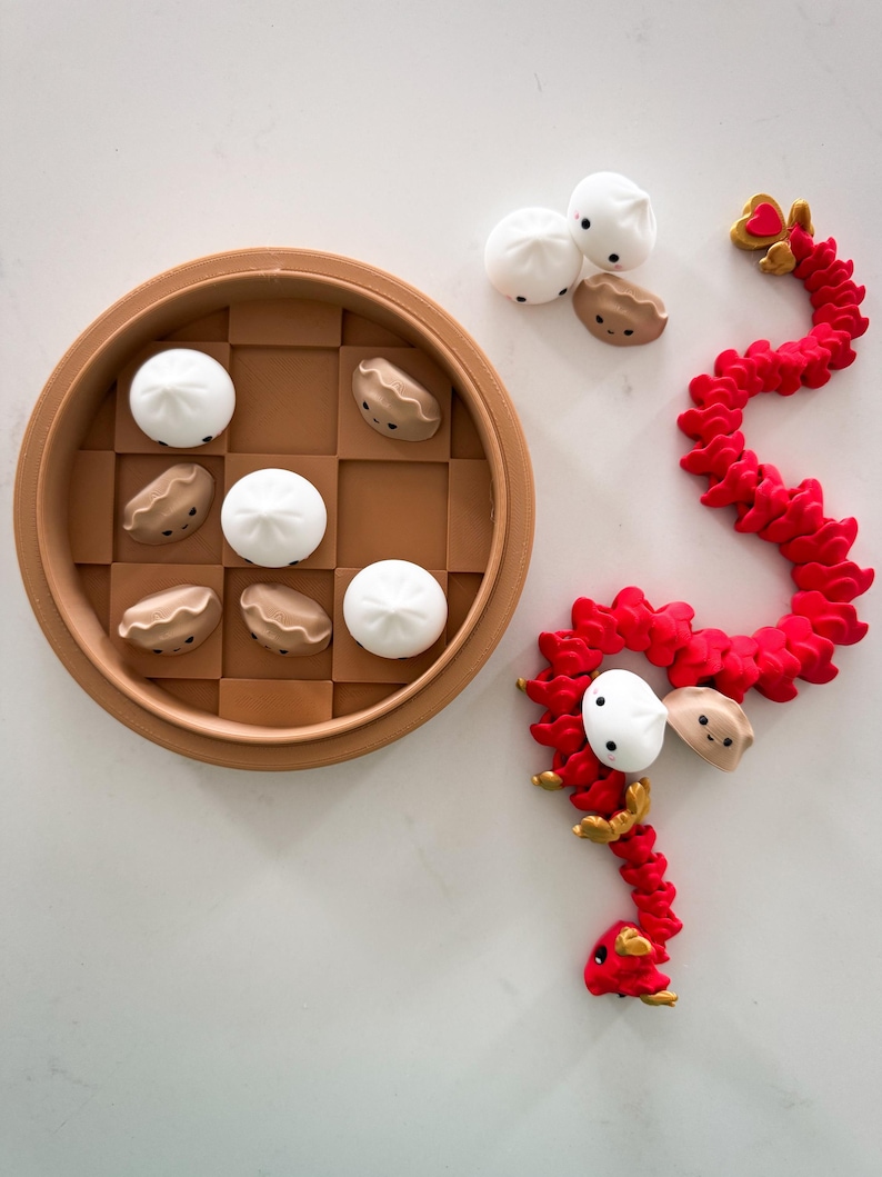 May include: A red dragon toy with a heart on its head, made of a soft, flexible material. The dragon is curled up around three white and brown steamed bun toys. The buns are also in a brown bamboo steamer.
