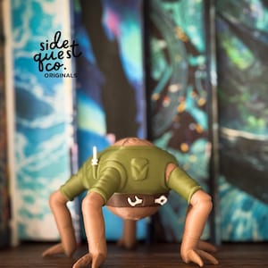 May include: A green and brown figurine of a creature with four limbs, a brown belt, and a green shirt. The figurine is on a wooden surface with colourful book spines in the background. The text "side quest co. originals" is visible.