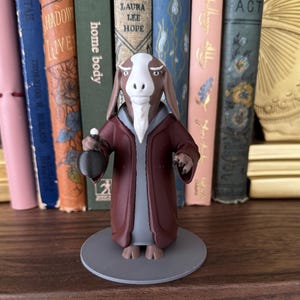 May include: A figurine of a goat-like creature wearing a burgundy robe and holding a small pouch. The figure has a white face and beard, standing on a gray circular base. The background features a bookshelf with colorful books.