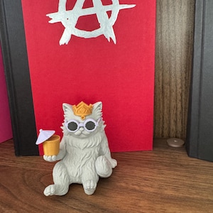 May include: A white cat figurine wearing a gold crown and white round sunglasses, holding a yellow drink with a small white umbrella. Behind the cat is a red book cover featuring a large, white, hand-drawn anarchy symbol centered on the front.