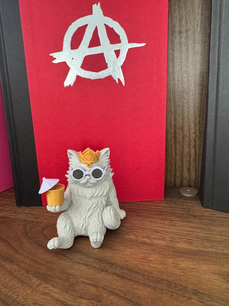 May include: A white cat figurine wearing a gold crown and white round sunglasses, holding a yellow drink with a small white umbrella. Behind the cat is a red book cover featuring a large, white, hand-drawn anarchy symbol centered on the front.