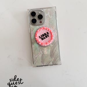 May include: Clear phone case with a pearlescent design and a pink and white pop socket. The pop socket has the words "LIBRA BABY" in black lettering. The phone case has a triple-lens camera.