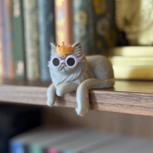 May include: A gray cat figurine wearing a gold crown and round sunglasses. The cat is lounging on a wooden shelf, with a blurred background of books and decorative items. The cat's tail hangs over the edge of the shelf.