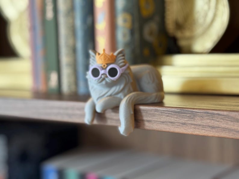 May include: A gray cat figurine wearing a gold crown and round sunglasses. The cat is lounging on a wooden shelf, with a blurred background of books and decorative items. The cat's tail hangs over the edge of the shelf.