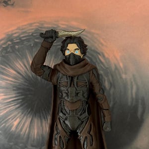 May include: A plastic action figure of a character with dark hair and bright blue eyes, wearing a black face mask and a brown and black textured suit with a cape. The figure holds a small, light-coloured curved blade raised above its head.