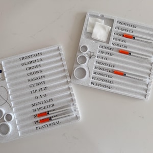 May include: Two white plastic trays with syringes and labels for cosmetic procedures. The labels include terms like "Frontalis," "Glabella," and "Masseter." Each tray has multiple syringes with orange plungers and a section for cotton pads.