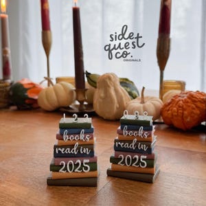 Può includere: Due candele a forma di libro con la scritta "books read in 2025" e numeri in cima. Le candele sono su un tavolo di legno con zucche, zucche e candele accese sullo sfondo. Il logo Side Quest Co. è visibile.