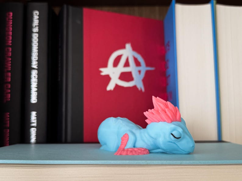 May include: A light blue fantasy creature figurine with pink details, resting on a shelf. The figurine has closed eyes and a relaxed posture. Behind it are books, including one with a red cover and a white symbol.