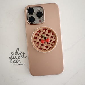 Könnte beinhalten: Ein roségoldenes Telefon mit einem Kirschkuchen-Telefonhalter. Der Telefonhalter ist rund mit einem Gitterkrustendesign und zwei roten Kirschen. Der Text "side quest co. ORIGINALS" ist sichtbar.