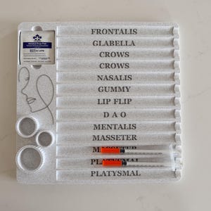 May include: A light gray plastic tray with labeled sections for facial treatments. The tray includes the text "FRONTALIS" to "PLATYSMAL" and has two syringes with orange plungers. Also included are three circular molds and an alcohol prep pad.