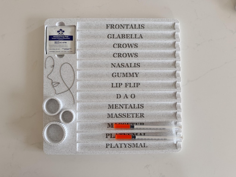 May include: A light gray plastic tray with labeled sections for facial treatments. The tray includes the text "FRONTALIS" to "PLATYSMAL" and has two syringes with orange plungers. Also included are three circular molds and an alcohol prep pad.