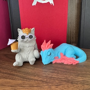 May include: A light gray cat figurine wearing a gold crown and white-rimmed sunglasses, holding a small yellow cup with a white umbrella, positioned next to a light blue dragon figurine with pink spiky hair and pink wing accents.