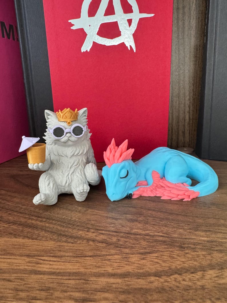 May include: A light gray cat figurine wearing a gold crown and white-rimmed sunglasses, holding a small yellow cup with a white umbrella, positioned next to a light blue dragon figurine with pink spiky hair and pink wing accents.