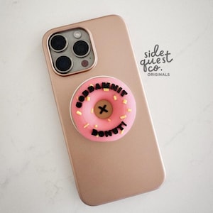 May include: A rose gold phone with a pink donut phone grip that says "GODDAMNIT DONUT" in black letters. The donut has yellow sprinkles and a black "X" in the centre. The phone has a triple-lens camera.