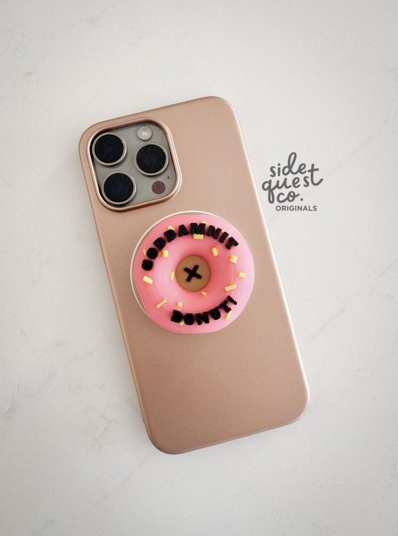May include: A rose gold phone with a pink donut phone grip that says "GODDAMNIT DONUT" in black letters. The donut has yellow sprinkles and a black "X" in the centre. The phone has a triple-lens camera.