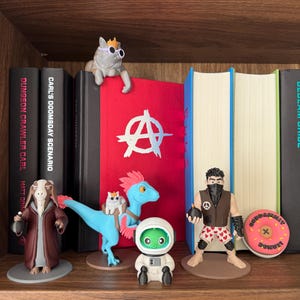 May include: A wooden shelf holds several figurines: a cat wearing sunglasses, a goat-like character in a robe, a blue dinosaur with a cat, an astronaut, and a muscular figure. A pink donut with text is also visible.