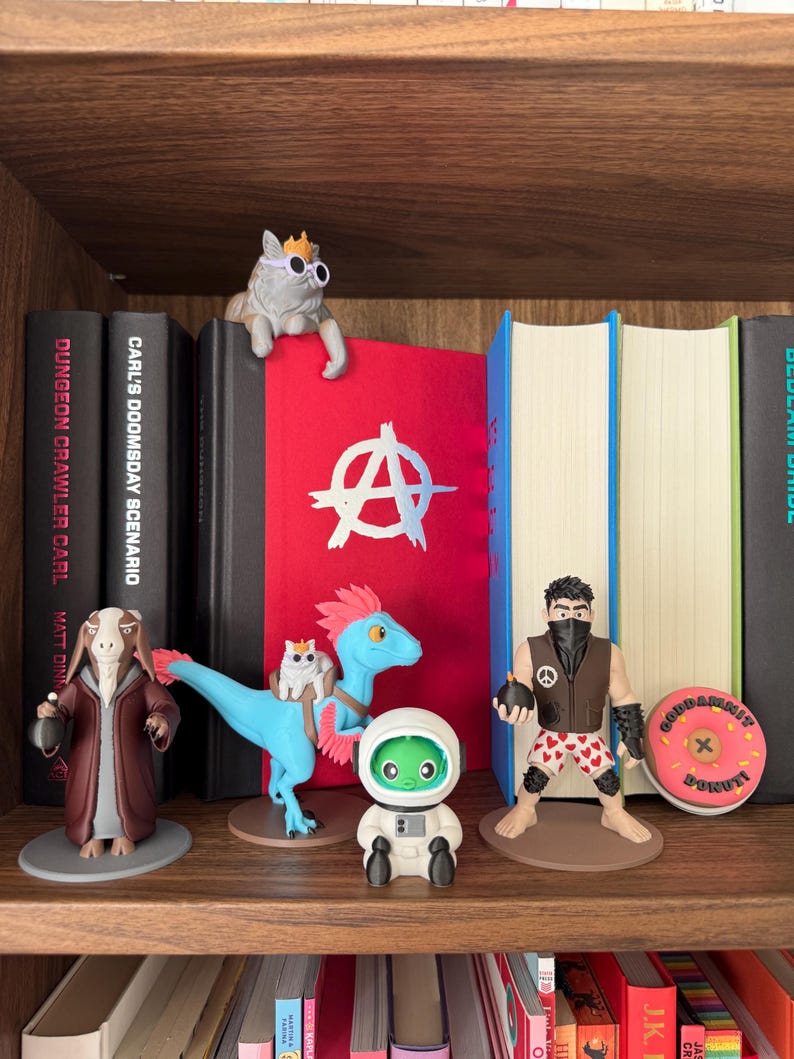 May include: A wooden shelf holds several figurines: a cat wearing sunglasses, a goat-like character in a robe, a blue dinosaur with a cat, an astronaut, and a muscular figure. A pink donut with text is also visible.