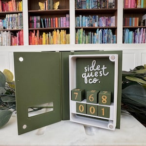 Puede incluir: Una caja en forma de libro verde oliva abierta, con un marco blanco y un interior verde oscuro. La caja presenta una cerradura de combinaci&oacute;n con los n&uacute;meros 768 y 001. El texto "side quest co." est&aacute; escrito en escritura blanca. Fondo con estanter&iacute;as.