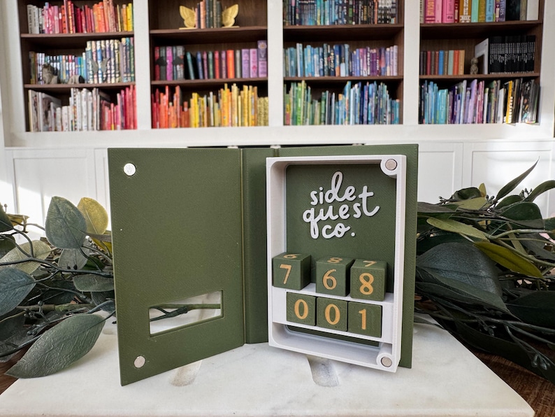 Puede incluir: Una caja en forma de libro verde oliva abierta, con un marco blanco y un interior verde oscuro. La caja presenta una cerradura de combinaci&oacute;n con los n&uacute;meros 768 y 001. El texto "side quest co." est&aacute; escrito en escritura blanca. Fondo con estanter&iacute;as.