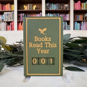 Puede incluir: Un contador de libros verde y dorado con el texto "Books Read This Year" y el n&uacute;mero "001" mostrado. El contador est&aacute; situado sobre un fondo de estanter&iacute;as llenas de libros y vegetaci&oacute;n.