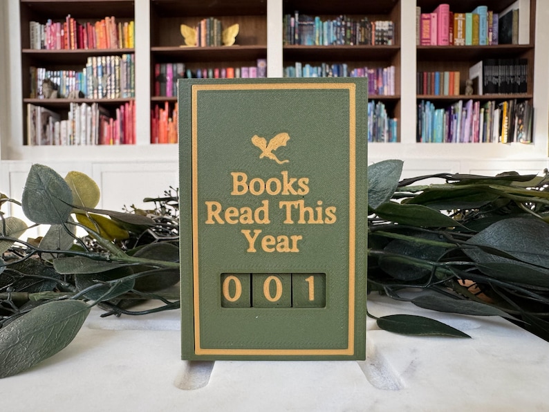 Puede incluir: Un contador de libros verde y dorado con el texto "Books Read This Year" y el n&uacute;mero "001" mostrado. El contador est&aacute; situado sobre un fondo de estanter&iacute;as llenas de libros y vegetaci&oacute;n.