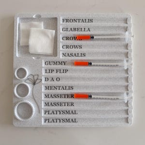 May include: A gray, textured tray with labeled sections for cosmetic procedures. Includes syringes with orange plungers, cotton pads, and circular molds. The tray is labeled with anatomical terms like "Frontalis" and "Glabella".