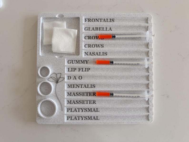 May include: A gray, textured tray with labeled sections for cosmetic procedures. Includes syringes with orange plungers, cotton pads, and circular molds. The tray is labeled with anatomical terms like "Frontalis" and "Glabella".