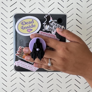 Op de afbeelding: Een zwarte popsocket met een silhouet van een figuur met een capuchon die een kopje vasthoudt, op een lichtpaarse achtergrond. De popsocket is bevestigd aan een zwart apparaat bedekt met stickers, waaronder een met "Dead Inside".
