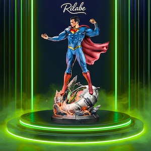 Puede incluir: Estatua de Superman en pose dinámica, vistiendo un traje azul con capa y botas rojas. La figura se alza sobre una estructura metálica dañada con llamas, sobre un fondo verde y negro. La palabra "Rilabe" está escrita en cursiva en la parte superior.