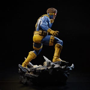 May include: A detailed figurine of a superhero in a dynamic pose. The figure is clad in a blue and yellow costume, with a red visor. The figure is standing on a rocky base, with a dark background.