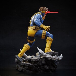 May include: A detailed figurine of a superhero in a dynamic pose, wearing a blue and yellow costume. The figure has a red beam emanating from its eyes, and is standing on a rocky base. The figure is approximately 12 inches tall.