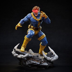 May include: A detailed figurine of a superhero in a dynamic pose. The figure is clad in a blue and yellow costume, with a red visor. The base is a rocky terrain with white accents, and the overall aesthetic is a comic book style.