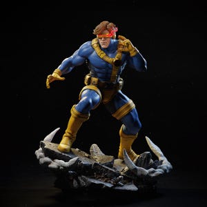 May include: A detailed figurine of Cyclops from the X-Men. The figure is in a dynamic pose, wearing a blue and yellow costume with a red visor. The base is a rocky terrain with snow-like accents. The figure is approximately 12 inches tall.