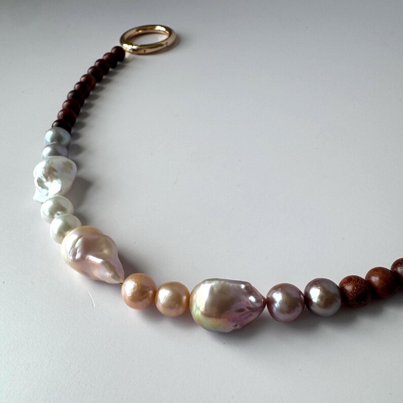 May include: A necklace featuring a mix of large, irregularly shaped pearls in shades of white, pink, and iridescent hues, interspersed with small, round, dark brown wooden beads. The necklace has a gold-colored ring clasp.