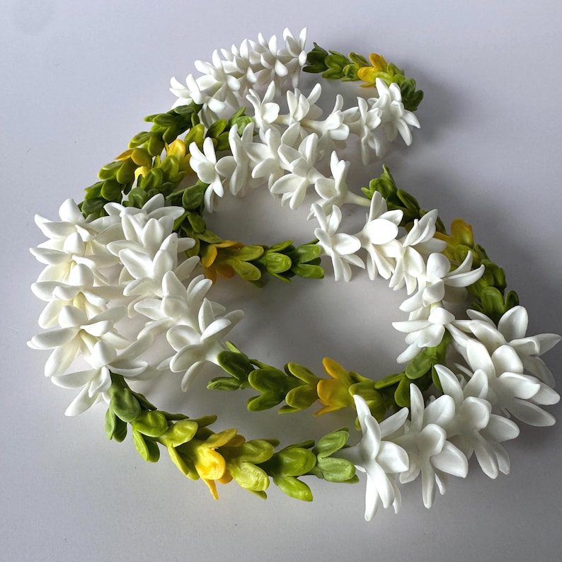 May include: A floral lei featuring white blossoms and green and yellow leaves. The lei is arranged in a circular shape, with the flowers and leaves evenly distributed around the circumference. The background is a solid white.