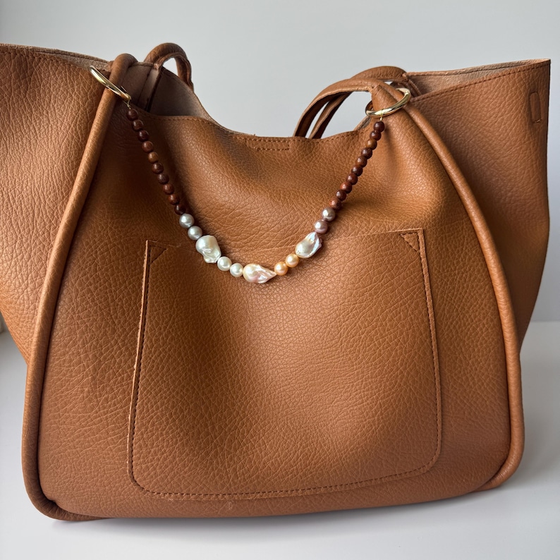 May include: A brown leather tote bag with a decorative pearl and wooden bead strap. The bag has a large pocket on the front and two handles. The pearl strap is attached to the bag with gold-colored rings.