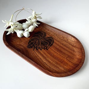 May include: A wooden oval tray with a dark brown finish, featuring a leaf design and the name "Ava" engraved on the surface. A pair of silver hoop earrings adorned with white flowers rests on the tray.