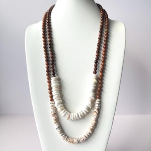 May include: Two layered necklace displayed on a white mannequin. The top layer features small, dark brown wooden beads. The bottom layer is made of white and tan shell-shaped beads, creating a natural, earthy aesthetic.