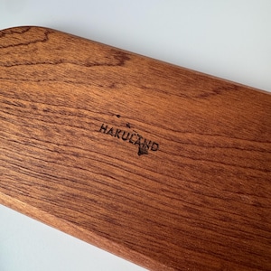 May include: A rectangular wooden serving board with a rich brown finish. The text "HAKULAND" is etched into the wood, along with a small island graphic. The board has a smooth, polished surface and a natural wood grain pattern.