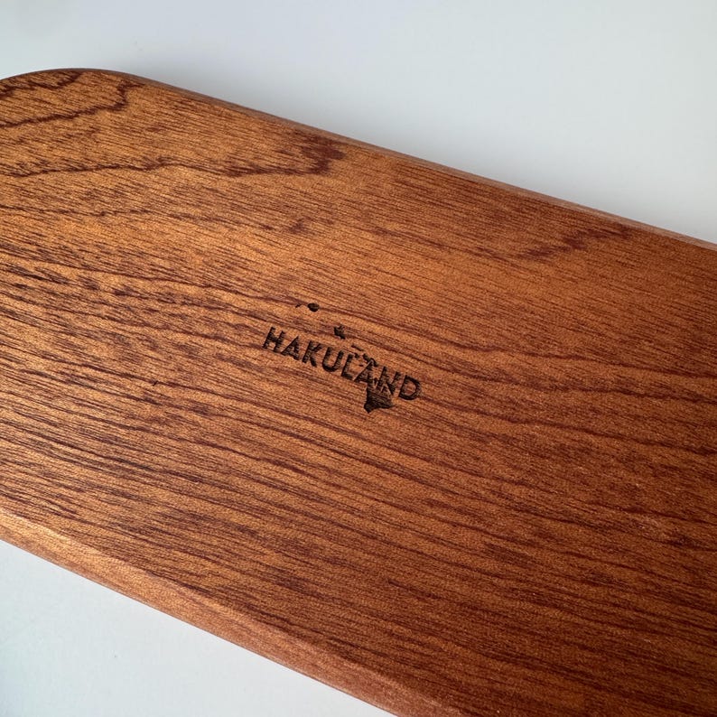 May include: A rectangular wooden serving board with a rich brown finish. The text "HAKULAND" is etched into the wood, along with a small island graphic. The board has a smooth, polished surface and a natural wood grain pattern.